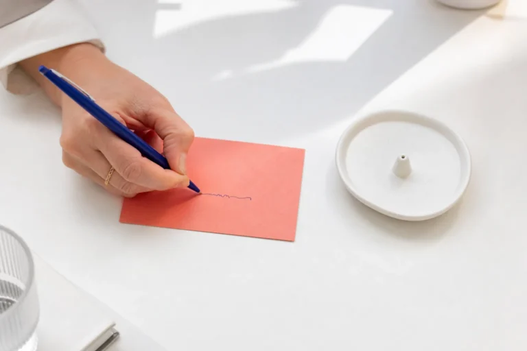 Hand schrijft één regel op een koraalkleurig kaartje naast een koude wierookhouder.